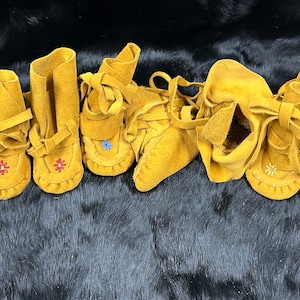 May include: A selection of mustard-yellow suede baby moccasins, handmade with decorative stitching and beaded details in red, blue, and white. Each pair has soft ties for fastening. The moccasins are displayed on a dark, textured surface.