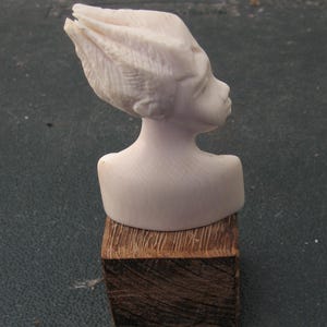 May include: A light-colored, carved bust of a head with stylized, upward-sweeping hair, positioned in profile. The sculpture features smooth facial contours and rests upon a dark, rectangular wooden base with a visible, textured grain pattern.