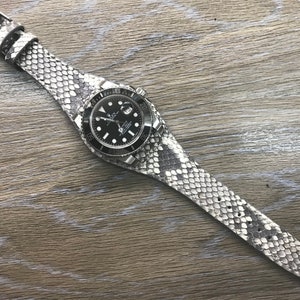 Puede incluir: Un reloj plateado con una esfera negra y una correa de piel de serpiente blanca.