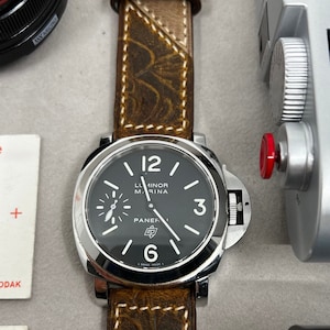 以下が含まれることがあります： 黒色の文字盤と茶色の革バンドが付いた腕時計。文字盤には白い目盛りと「Luminor Marina」と「Panerai」の文字が書かれている。