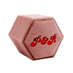 May include: A pink velvet hexagonal ring box with the letters "P&R" in red script. The box is closed and has a soft, plush appearance. The design is simple and elegant, suitable for jewelry.