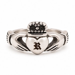 May include: A silver-toned Claddagh ring featuring two hands holding a central heart topped with a crown. The heart is engraved with the letter R in a gothic-style font. The metal has a polished finish with darkened recessed details.
