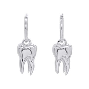 Puede incluir: Un par de pendientes de aro plateados, cada uno con un dije de molar detallado. Los dijes de molar tienen una forma realista con raíces y detalles de esmalte visibles. Los pendientes se muestran sobre un fondo blanco.