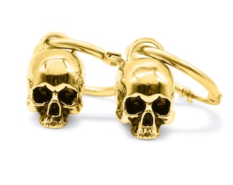 May include: A pair of gold-toned hoop earrings, each featuring a polished, three-dimensional skull charm suspended from a circular hoop. The skulls have detailed eye sockets, nasal cavities, and teeth, with a reflective, smooth metallic finish.