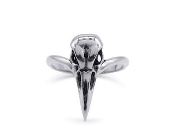 Puede incluir: Un anillo plateado con un diseño de calavera de cuervo. El anillo presenta una representación detallada de un cráneo de ave, con un pico largo y puntiagudo y detalles intrincados. La banda se curva hacia los lados del cráneo.