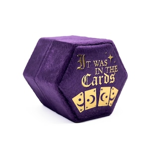 May include: A hexagonal, purple velvet box with gold lettering that reads "It Was In The Cards." Three gold playing cards with crescent moon designs are below the text. The box is on a white background.