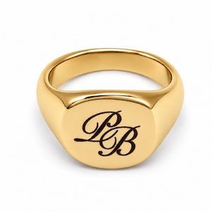 May include: A polished gold signet ring featuring a smooth, rounded band and a flat, circular face. The centre of the ring is engraved with the stylised, interconnected cursive letters P and B in a dark, contrasting tone against the gold surface.