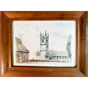 May include: A framed watercolor painting depicting a stone church with a tall square tower and a separate stone building to the left. The scene includes a low stone wall, trees, and a pale blue sky. The artwork is enclosed in a light brown wooden frame.