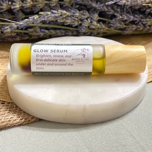 Op de afbeelding: Een klein glazen flesje met een houten dop, gevuld met een heldere vloeistof. Het flesje is gelabeld met "Glow Serum" en heeft de tekst "Brighten, renew, and firm delicate skin under and around the eyes." Het flesje staat op een wit rond oppervlak.