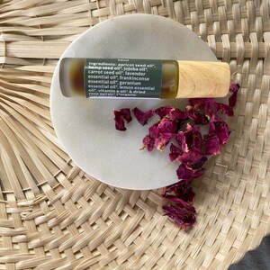 Op de afbeelding: Een 10 ml amberkleurige glazen rollerfles met een houten dop, gevuld met een gouden olie. De fles staat op een wit rond oppervlak met gedroogde rozenblaadjes. Het etiket op de fles vermeldt de ingredi&euml;nten: abrikozenpitolie, hennepzaadolie, jojoba-olie, wortelolie, lavendelolie, wierookolie, geraniumolie, citroenolie, vitamine E-olie en gedroogde rozenblaadjes. Het etiket vermeldt ook dat de ingredi&euml;nten biologisch zijn.