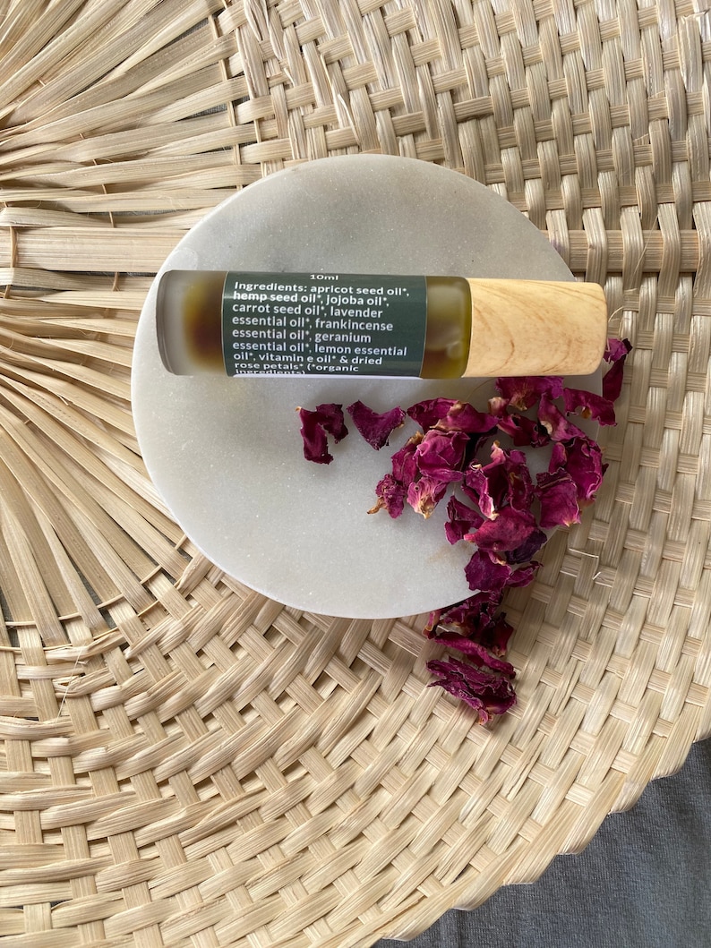 Op de afbeelding: Een 10 ml amberkleurige glazen rollerfles met een houten dop, gevuld met een gouden olie. De fles staat op een wit rond oppervlak met gedroogde rozenblaadjes. Het etiket op de fles vermeldt de ingredi&euml;nten: abrikozenpitolie, hennepzaadolie, jojoba-olie, wortelolie, lavendelolie, wierookolie, geraniumolie, citroenolie, vitamine E-olie en gedroogde rozenblaadjes. Het etiket vermeldt ook dat de ingredi&euml;nten biologisch zijn.