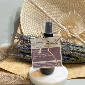 May include: A 2 ounce bottle of Just Breathe Spritz, a natural aromatherapy spray for clarity, focus, and release. The bottle is brown glass with a black spray top and is tied with twine to a small card with the product name and ingredients. The ingredients are eucalyptus radiata, lavender, frankincense, orange, rosemary, spearmint, and distilled water.