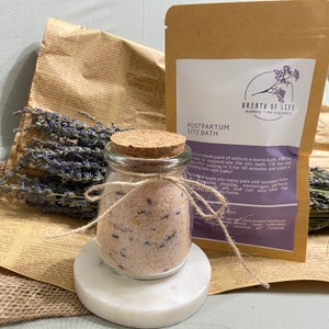 May include: A glass jar filled with pink bath salts with lavender buds and a cork lid. The jar is tied with twine and sits on a white surface. The label on the bag reads "Postpartum Sitz Bath" and "Breath of Life mommy + me organics".
