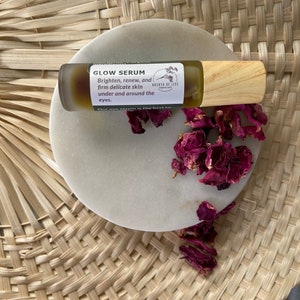 Op de afbeelding: Een klein glazen flesje met een houten dop en een etiket met de tekst "GLOW SERUM" en "Brighten, renew, and firm delicate skin under and around the eyes." Het flesje staat op een wit marmeren oppervlak met gedroogde rozenblaadjes.