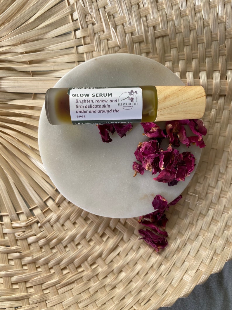 Op de afbeelding: Een klein glazen flesje met een houten dop en een etiket met de tekst "GLOW SERUM" en "Brighten, renew, and firm delicate skin under and around the eyes." Het flesje staat op een wit marmeren oppervlak met gedroogde rozenblaadjes.