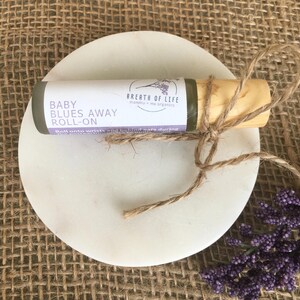 May include: A small, green glass bottle with a wooden cap and a brown twine bow. The bottle is labeled "Baby Blues Away Roll-On" and "Breath of Life mommy + me organics".