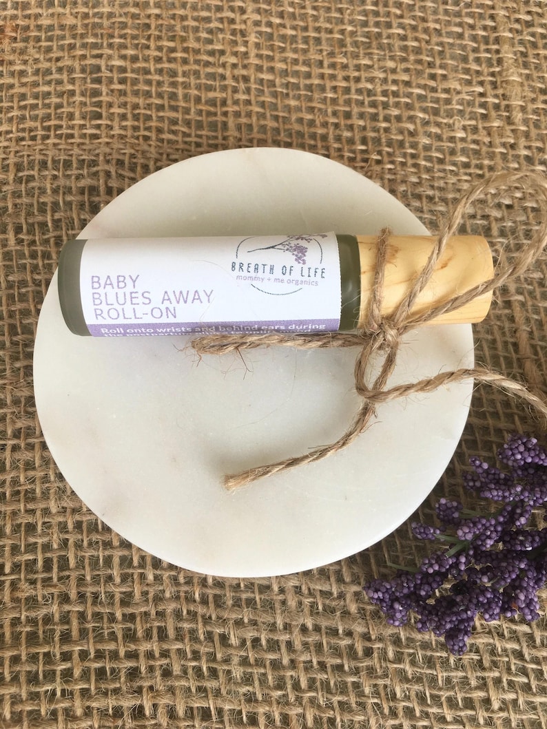 May include: A small, green glass bottle with a wooden cap and a brown twine bow. The bottle is labeled "Baby Blues Away Roll-On" and "Breath of Life mommy + me organics".