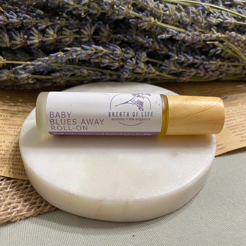 May include: A small glass bottle with a wooden roller ball top. The bottle is labeled "Baby Blues Away Roll-On" and "Breath of Life mommy + me organics".
