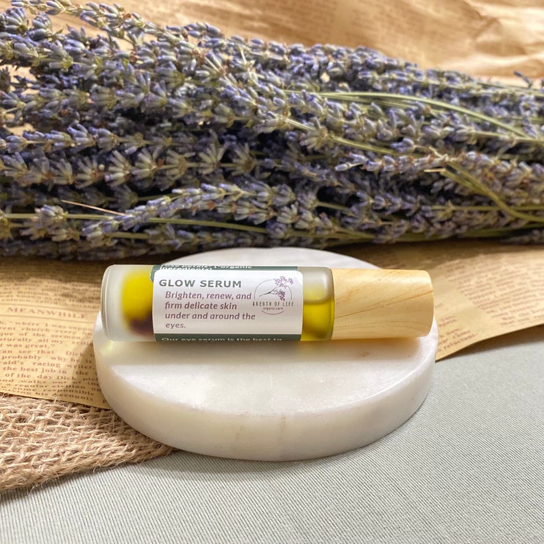 Op de afbeelding: Een klein glazen flesje met een houten dop, gevuld met een heldere vloeistof. Het flesje is gelabeld met "Glow Serum" en heeft de tekst "Brighten, renew, and firm delicate skin under and around the eyes." Het flesje staat op een wit rond oppervlak.