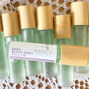 May include: A set of eight glass roller bottles with bamboo lids. The bottles are filled with a light green liquid. The label on one bottle reads "Baby Blues Away Roll On" and "Breath of Life mommy + me organics".