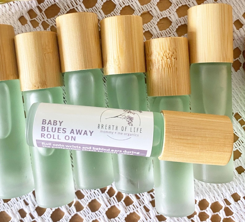 May include: A set of eight glass roller bottles with bamboo lids. The bottles are filled with a light green liquid. The label on one bottle reads "Baby Blues Away Roll On" and "Breath of Life mommy + me organics".
