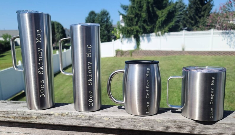 May include: Four stainless steel travel mugs with different sizes and inscriptions. The mugs are on a wooden surface. The largest mug is 30oz Skinny Mug, the next is 20oz Skinny Mug, the third is 14oz Coffee Mug, and the smallest is 14oz Camper Mug.