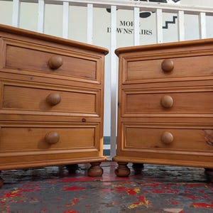 May include: A pair of matching light brown wooden bedside cabinets, each featuring three drawers with rounded wooden knobs and supported by four bun feet. The rectangular units have a smooth finish and a simple frame design on each drawer front.