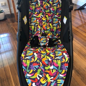 Könnte beinhalten: Ein schwarzer Kinderwagen mit einer bunten Sitzbezug. Das Muster zeigt abstrakte Formen in Blautönen, Grün, Gelb, Orange, Rot und Lila auf einem dunkelvioletten Hintergrund.