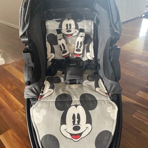 May include: A black stroller with a gray Mickey Mouse themed liner. The liner features a large Mickey Mouse face in the center and smaller Mickey Mouse ears and heads around the edges.