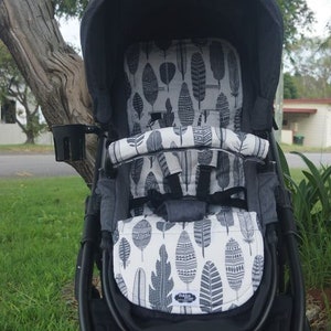 Pode incluir: Um carrinho de bebê preto com um forro de assento estampado em branco e cinza. O forro apresenta um design de penas.
