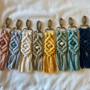 May include: Eight colorful macrame keychains with a small lava rock bead in the center of each. The keychains are made with a variety of colors, including pink, blue, white, green, yellow, and black.