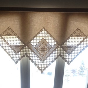 May include: A brown valance with a white crocheted design. The valance is hung over a window with three panes.