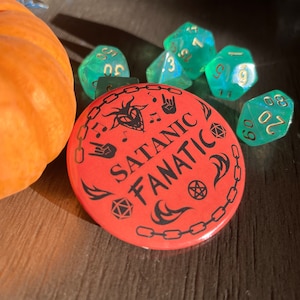 Puede incluir: Un botón rojo con un borde de cadena negro y el texto "SATANIC FANATIC" en negro. El botón tiene una ilustración en blanco y negro de una cabeza de diablo con cuernos, un corazón y notas musicales. También hay ilustraciones en blanco y negro de un pentagrama, una mano con un signo de rock and roll y llamas.