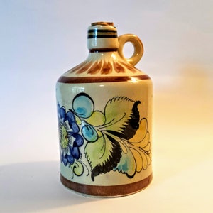 May include: A ceramic jug with a handle and a cork stopper. The jug is beige with brown accents and a floral design in blue, yellow, and green. The jug is approximately 6 inches tall.