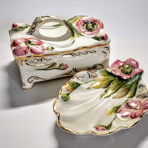 May include: A white porcelain trinket box with a floral design and gold accents. The box has a hinged lid with a handle and a matching dish with a scalloped edge.