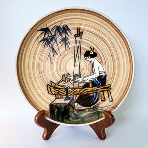 May include: Decorative ceramic plate with a hand-painted scene of a woman weaving. The plate has a light brown, wood-grain-like background with a white rim. The woman is depicted in black and blue clothing, with a yellow flower in her hair.