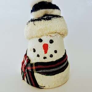 May include: A snowman figurine with a cream-colored body, black dot eyes, and a carrot nose. It wears a striped blue and white hat with a white pom-pom and a plaid scarf. A festive holiday decoration.