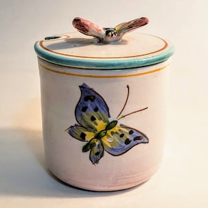 May include: A ceramic jar with a lid, featuring a hand-painted butterfly design. The jar is cylindrical, with a light pink base, a blue and yellow butterfly, and a lid topped with a butterfly figurine. The lid has a turquoise rim.
