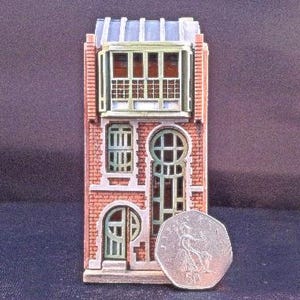 May include: A miniature model of a three-story brick building with a green roof and windows. The building has a door on the ground floor and a balcony on the second floor.