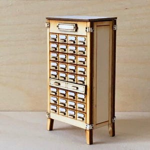 May include: A miniature wooden cabinet with numerous small drawers, ideal for storing small items. The cabinet has a light wood finish with darker wood trim and metal accents. The drawers have small white labels. The cabinet is a charming piece of decor.