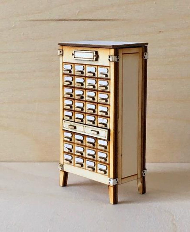 May include: A miniature wooden cabinet with numerous small drawers, ideal for storing small items. The cabinet has a light wood finish with darker wood trim and metal accents. The drawers have small white labels. The cabinet is a charming piece of decor.