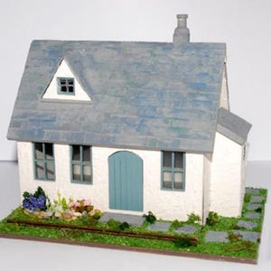May include: A white miniature cottage with a blue roof, blue door, and three windows. The cottage is surrounded by a small garden with green grass, flowers, and stone pathways.