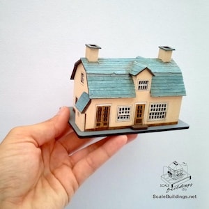 May include: A miniature model house with a light peach exterior, a blue-green shingled roof, and two chimneys. The house has multiple windows and a small door. The model is held in a hand and sits on a gray base.