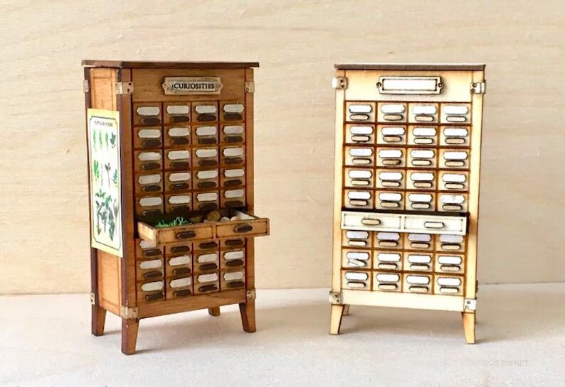 May include: Two miniature wooden apothecary cabinets. One is stained brown with the word "CURISOTIES" on top, and the other is unstained. Both have numerous small drawers with label holders and are on small legs.