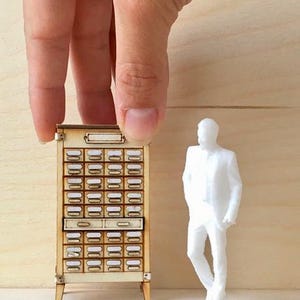 May include: Miniature wooden cabinet with numerous small drawers, each with a label holder. A small white figurine of a person stands nearby. The cabinet is held by a hand against a light wood background.