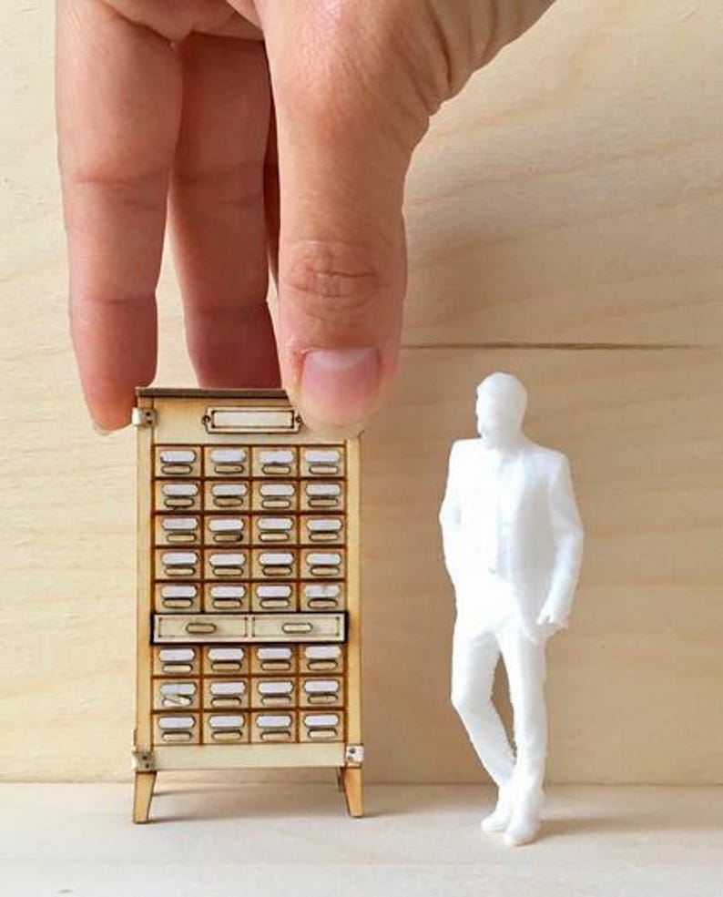 May include: Miniature wooden cabinet with numerous small drawers, each with a label holder. A small white figurine of a person stands nearby. The cabinet is held by a hand against a light wood background.