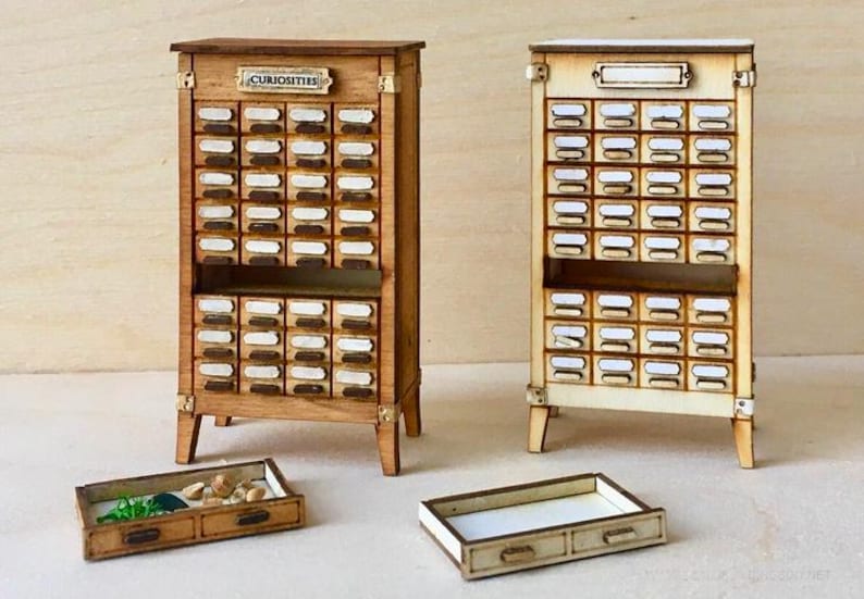 May include: Two miniature wooden cabinets with numerous small drawers, labeled with white tags. One cabinet is stained a darker brown, the other is lighter. The word "CURIOSITIES" is displayed on the darker cabinet. Two small trays are in front of the cabinets.