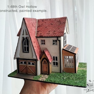 May include: A miniature Owl Hollow house model with a red tile roof, beige walls, and brown trim. The model includes a garage, windows, and a small porch. The base is green, and the text "1:48th Owl Hollow constructed, painted example" is visible.
