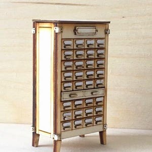 May include: A miniature wooden cabinet with numerous small drawers, ideal for organizing small items. The cabinet has a light wood finish with darker edges and metal corner accents. It features a rectangular top and is supported by four small legs.