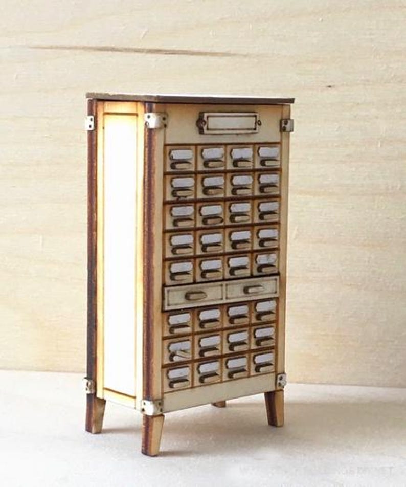 May include: A miniature wooden cabinet with numerous small drawers, ideal for organizing small items. The cabinet has a light wood finish with darker edges and metal corner accents. It features a rectangular top and is supported by four small legs.